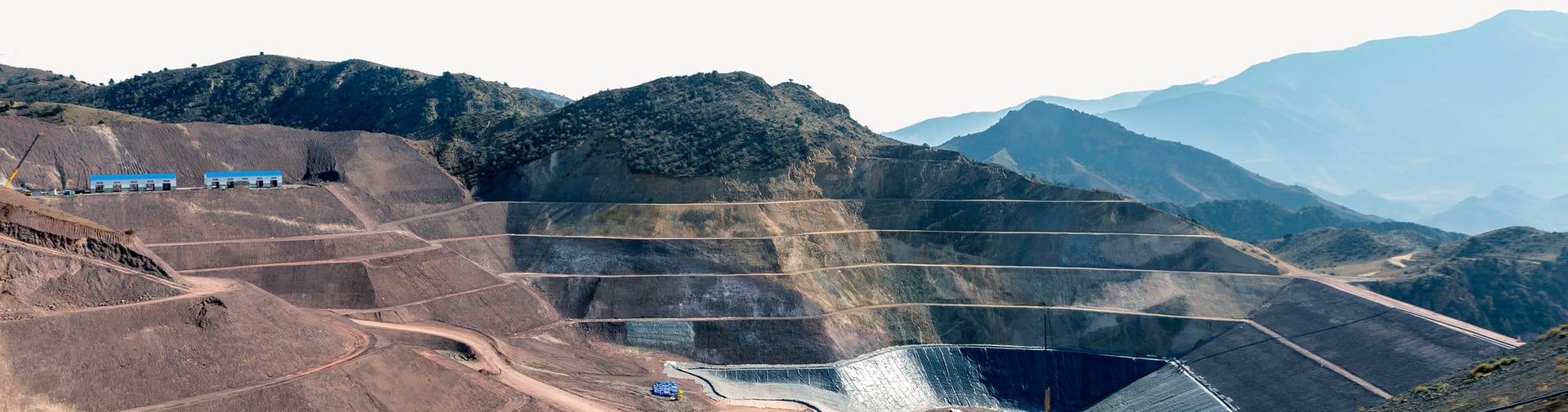 A landscape image of an open pit mine, in section, displaying mine tiers, forestry and mountains. A landscape image of an open pit mine, in section, displaying mine tiers, forestry and mountains.